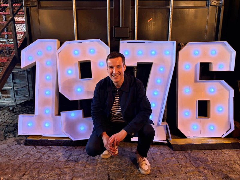 Laurence at his joint 50th party crouched in front of a set of large, white, illuminated numbers that make up the year 1976. Laurence is dressed in jeans, blue jacket and striped top, and smiling.